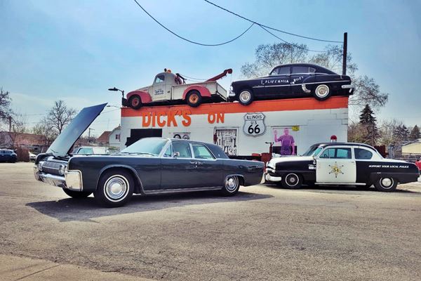 towing garage building, white with orange strip along parapet; tow truck and vintage car on roof, vintage cop car and 1960s car in front. DICKS ON 66 written in orange, US 66 shield