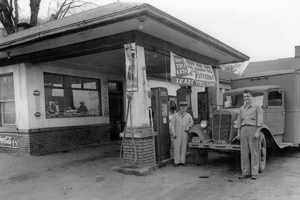 black and white 1940s, two men by truck in a gas station with pumps under hip roof