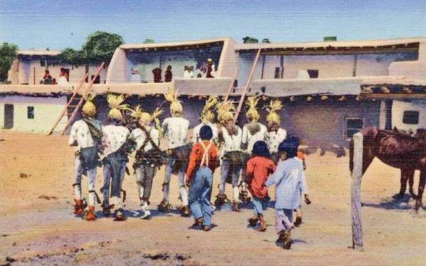 people in a single file dancing in a color 1940s postcard, adobe pueblo 2 story bldgs. with ladders behind them